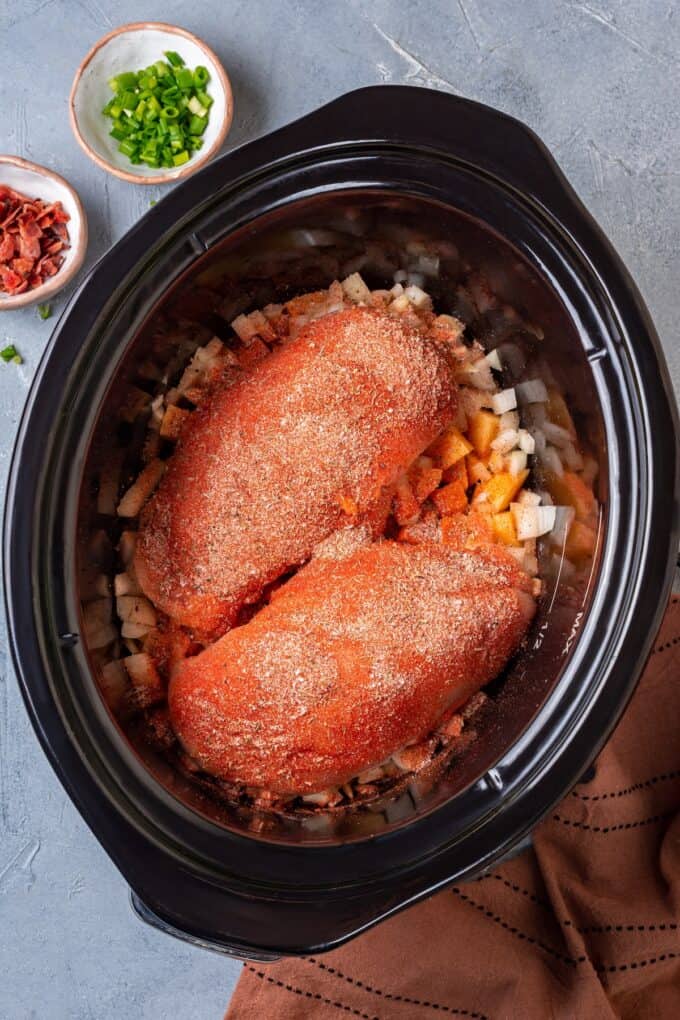Seasoned chicken breasts on top of chopped onions and potatoes in a slow cooker insert.
