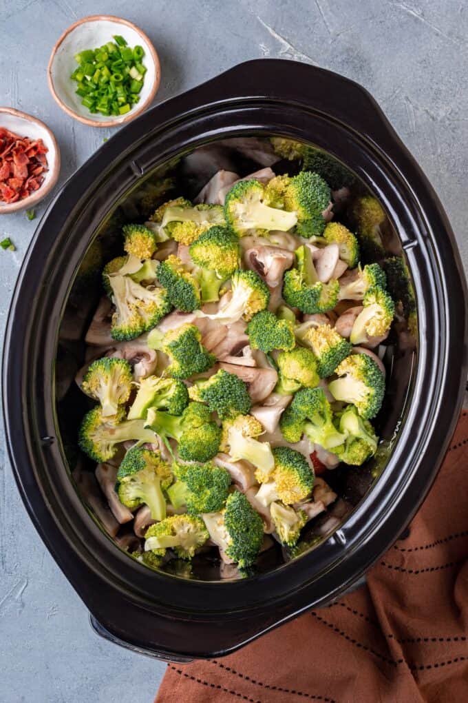 Mushrooms and broccoli being layered into a slow cooker insert.