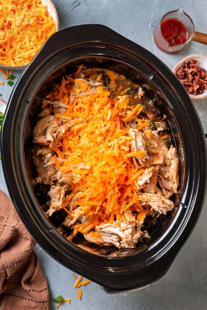 Shredded cheddar cheese being added on top of shredded chicken and potatoes in a slow cooker.