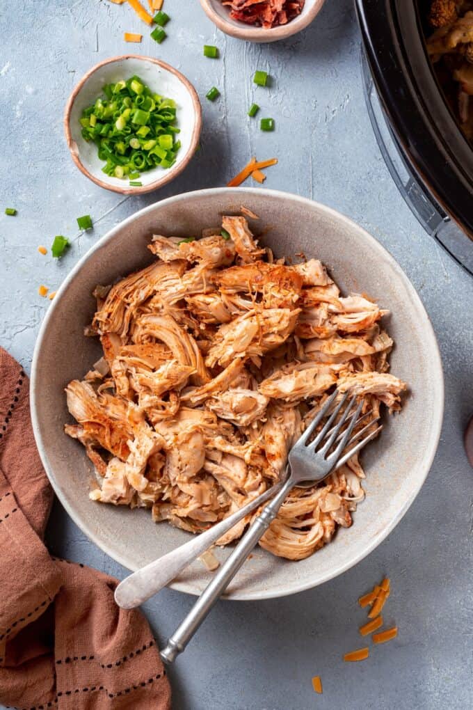 Shredded cooked chicken in a bowl with two forks to the side and a bowl of green onions in the back.
