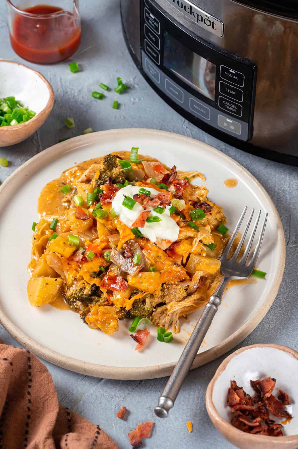 A dinner plate with a chicken and potato casserole with cheese, sour cream, green onions and bacon on top and a fork to the side with a crock pot in the background.