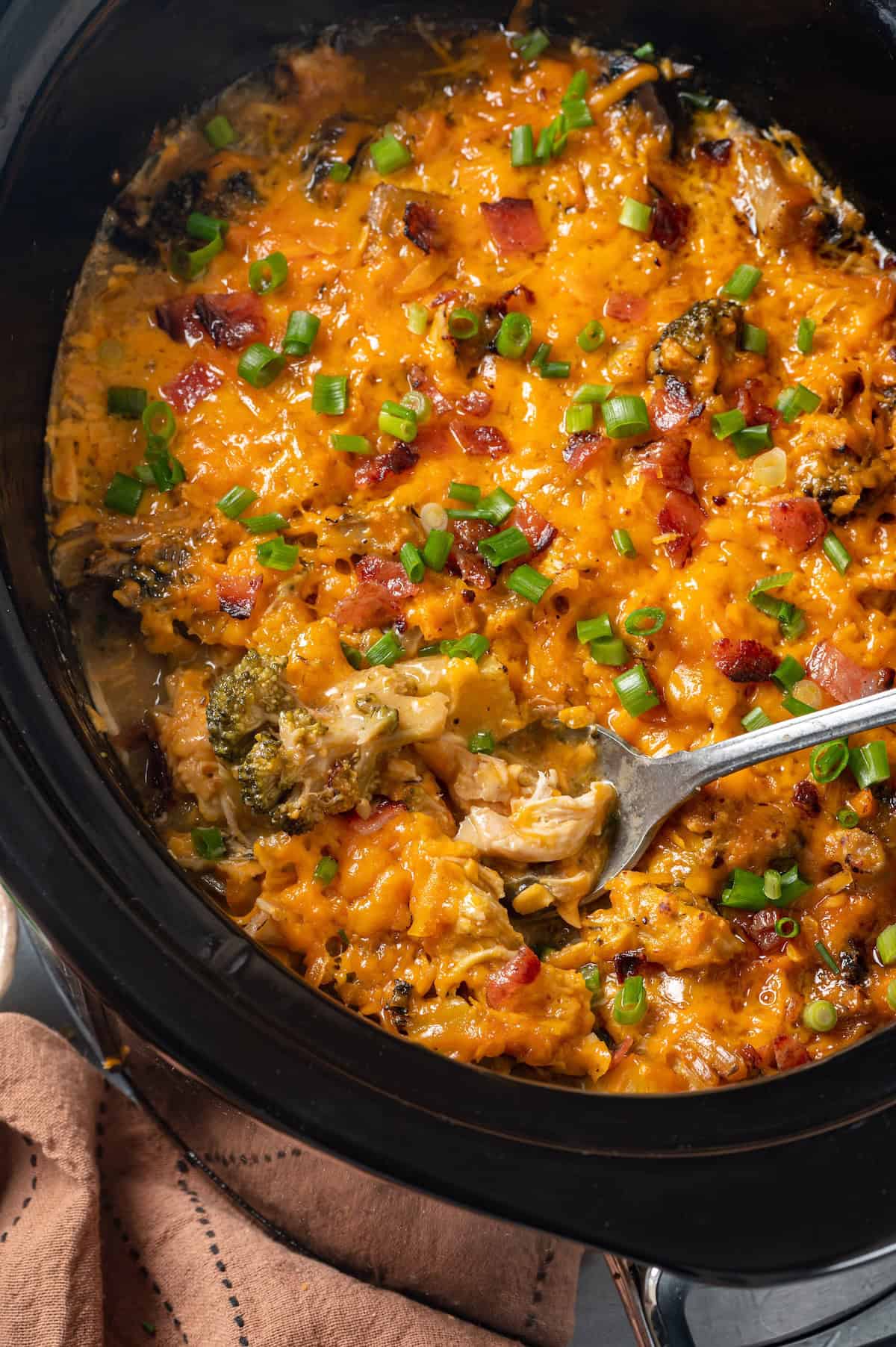 A serving spoon scooping up some cheesy chicken casserole with potatoes and broccoli from a slow cooker insert.