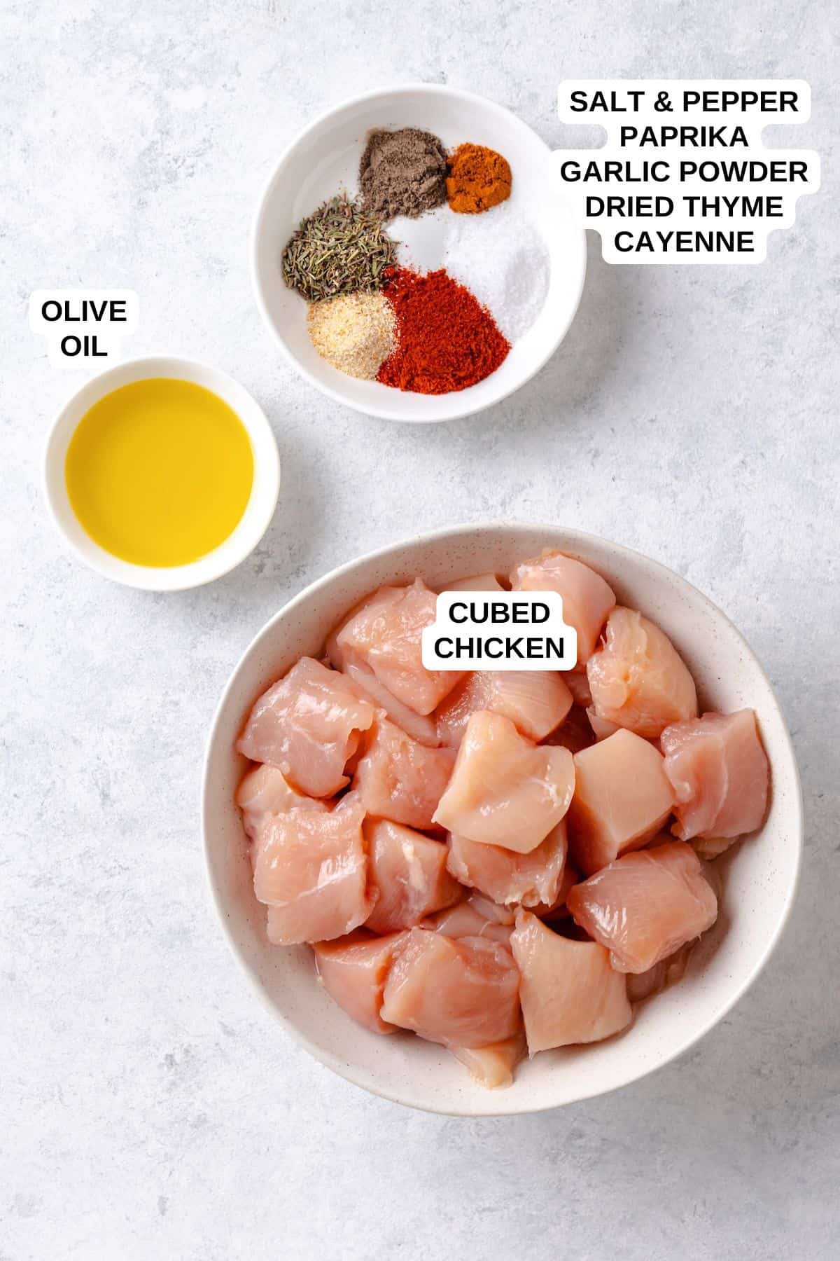 A bowl of cubed chicken breasts alongside bowls of olive oil and an arrangement of various spices.