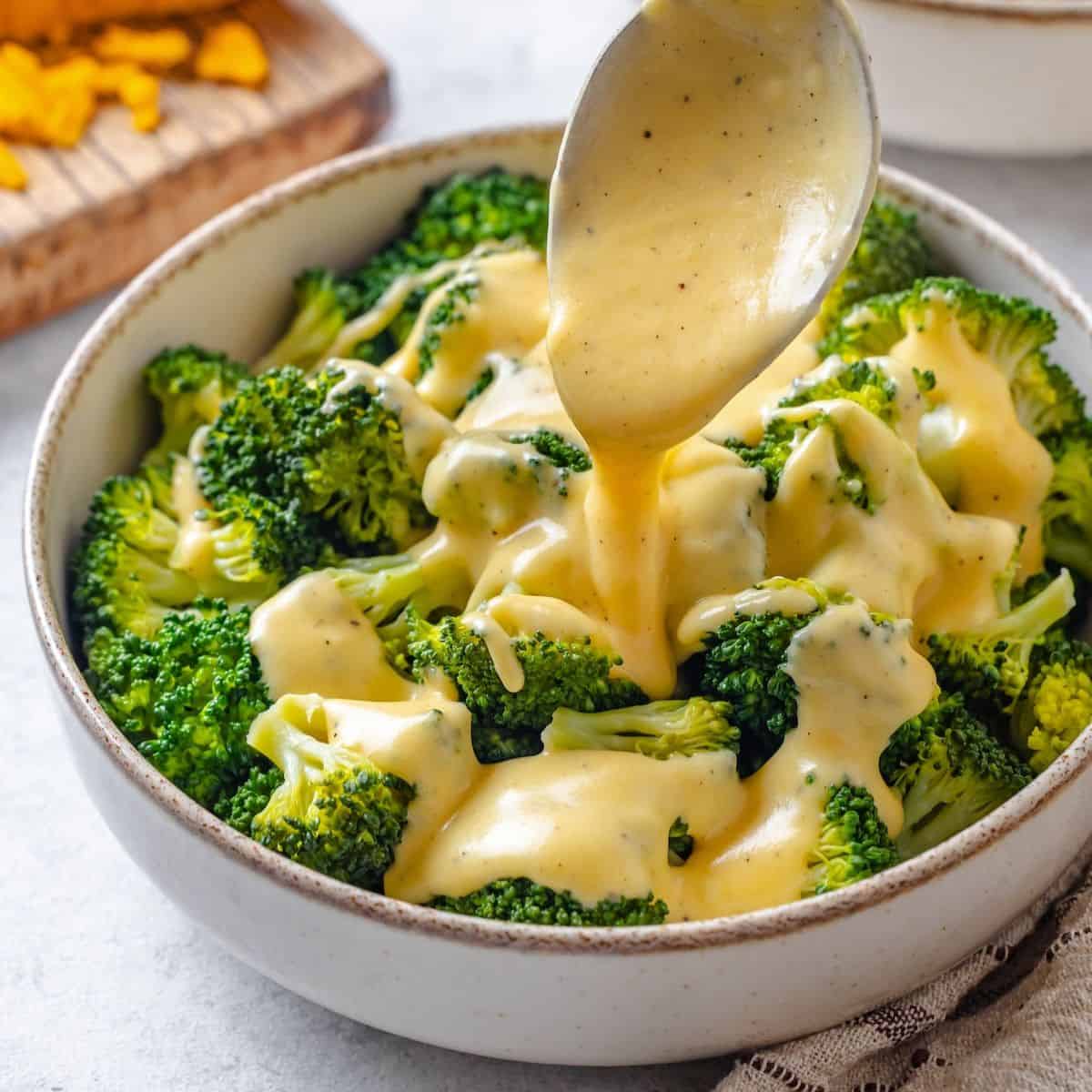 Close up of a spoon drizzling a cheese sauce over a bowl of steamed broccoli.