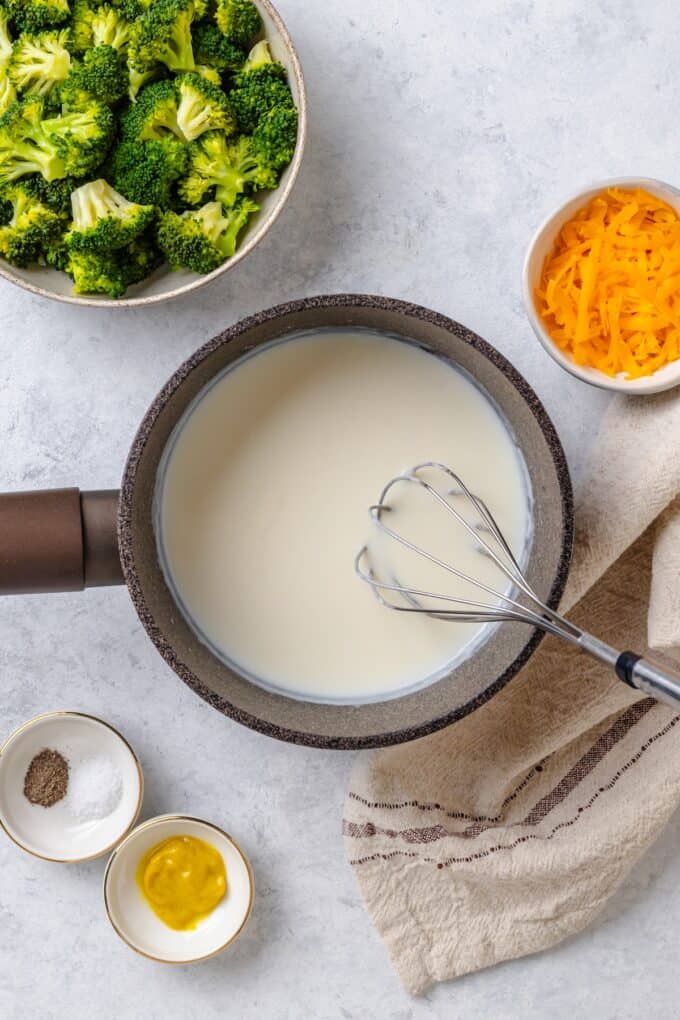 A whisk stirring milk into a roux in a small pot for creating a cheese sauce.