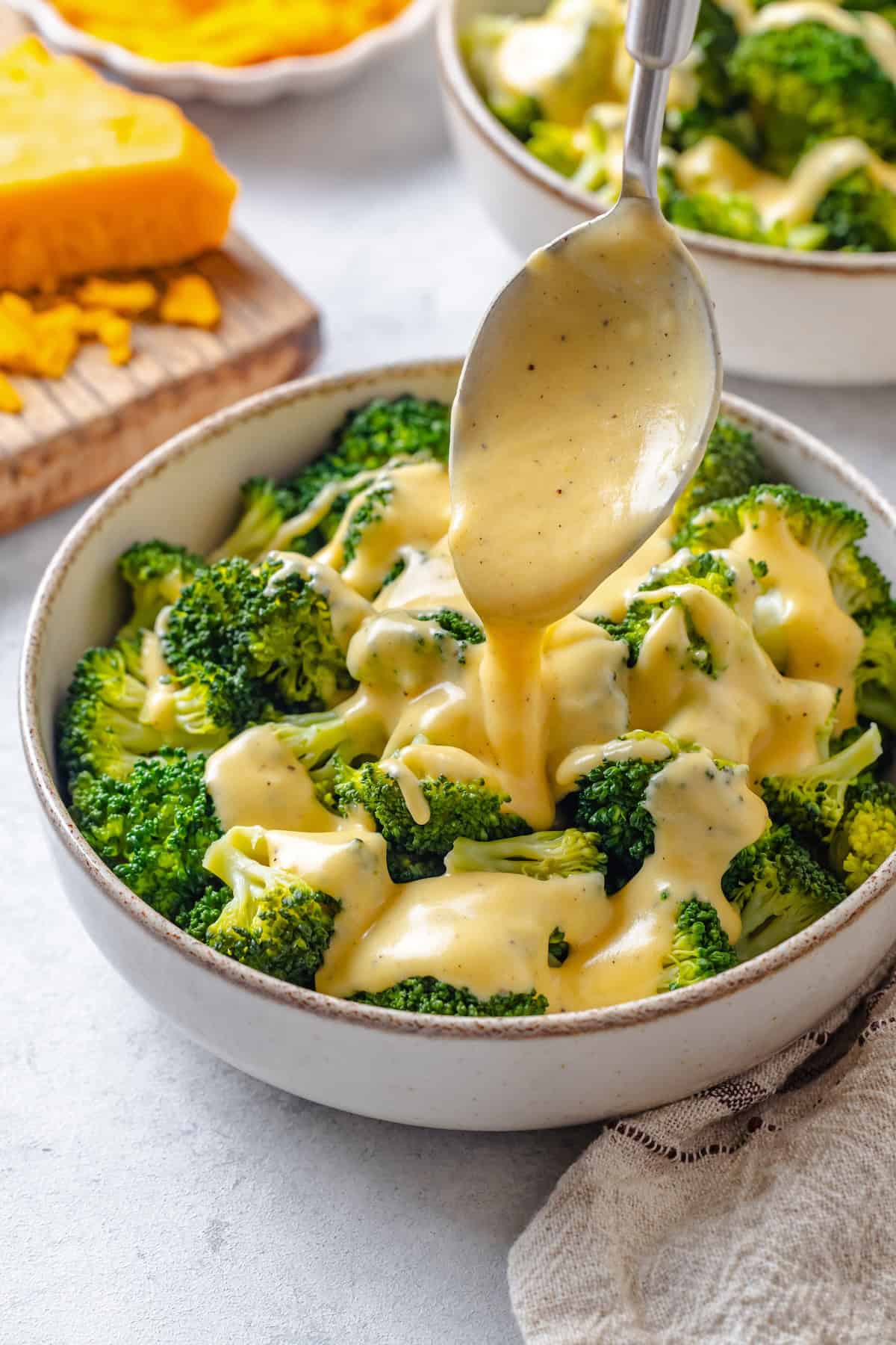 A spoon drizzling a cheese sauce over a bowl of steamed broccoli.