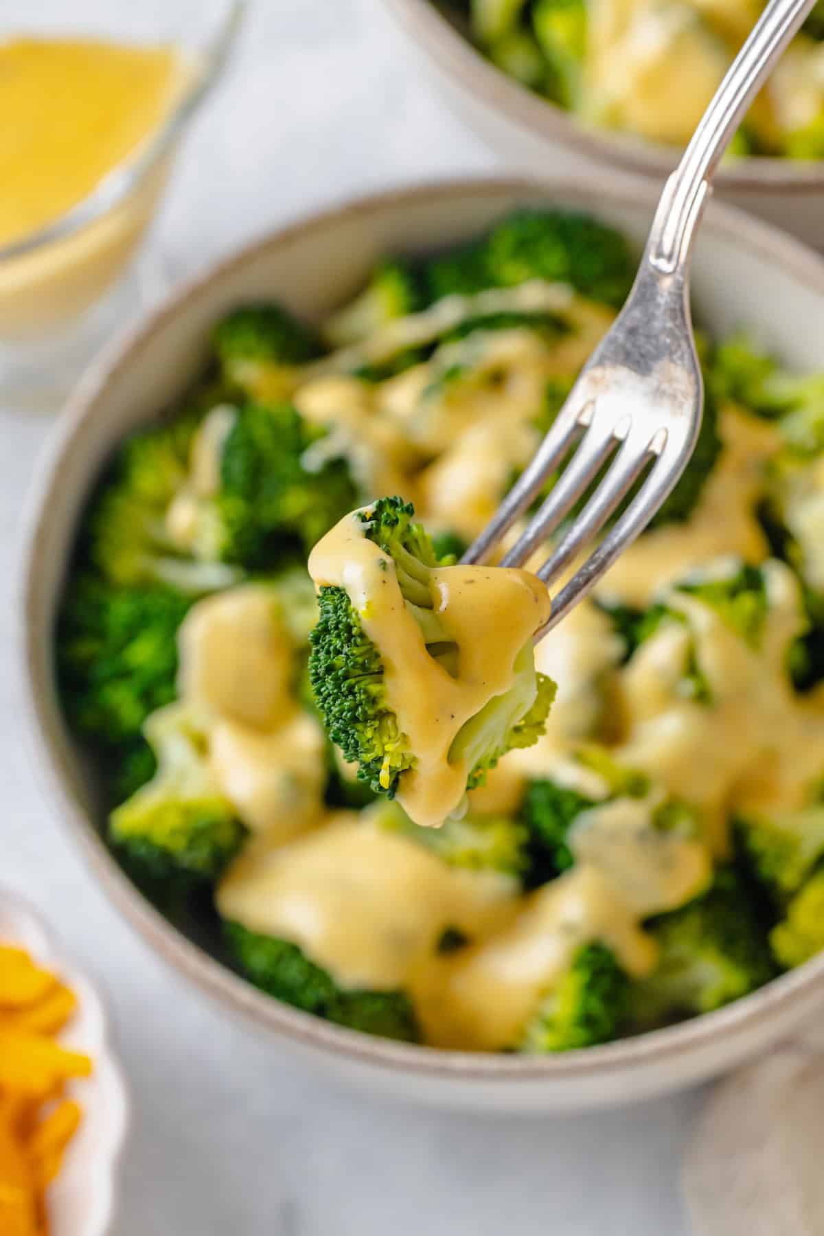 A fork lifting up a piece of steamed broccoli drizzled with a cheese sauce from a bowl.