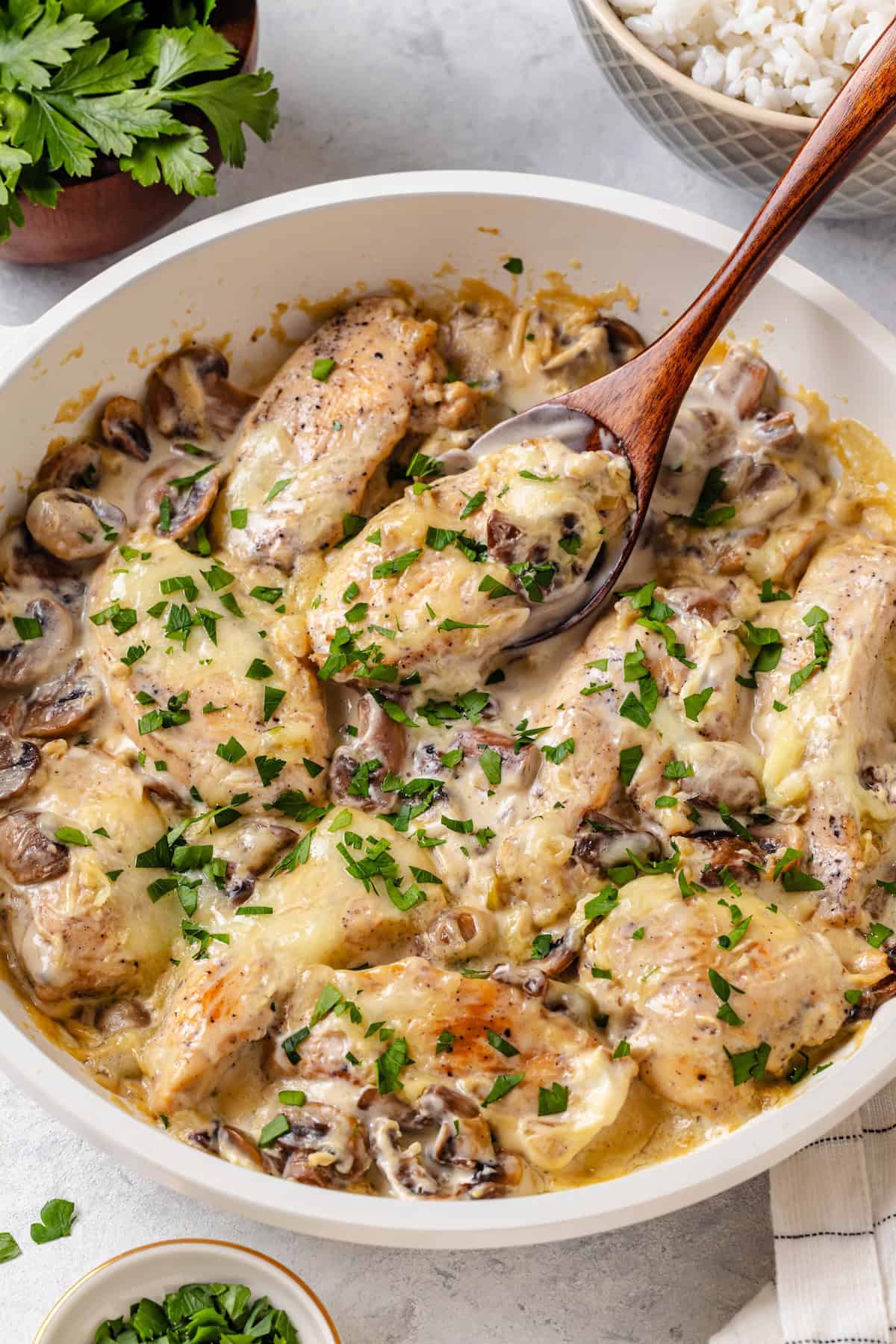 A spoon scooping up a piece of chicken from a skillet full of chicken in a cream cheese sauce with parsley sprinkled on top.