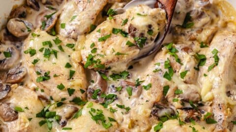 Close up of a spoon scooping up a piece of chicken from a skillet full of chicken in a cream cheese sauce with parsley sprinkled on top.