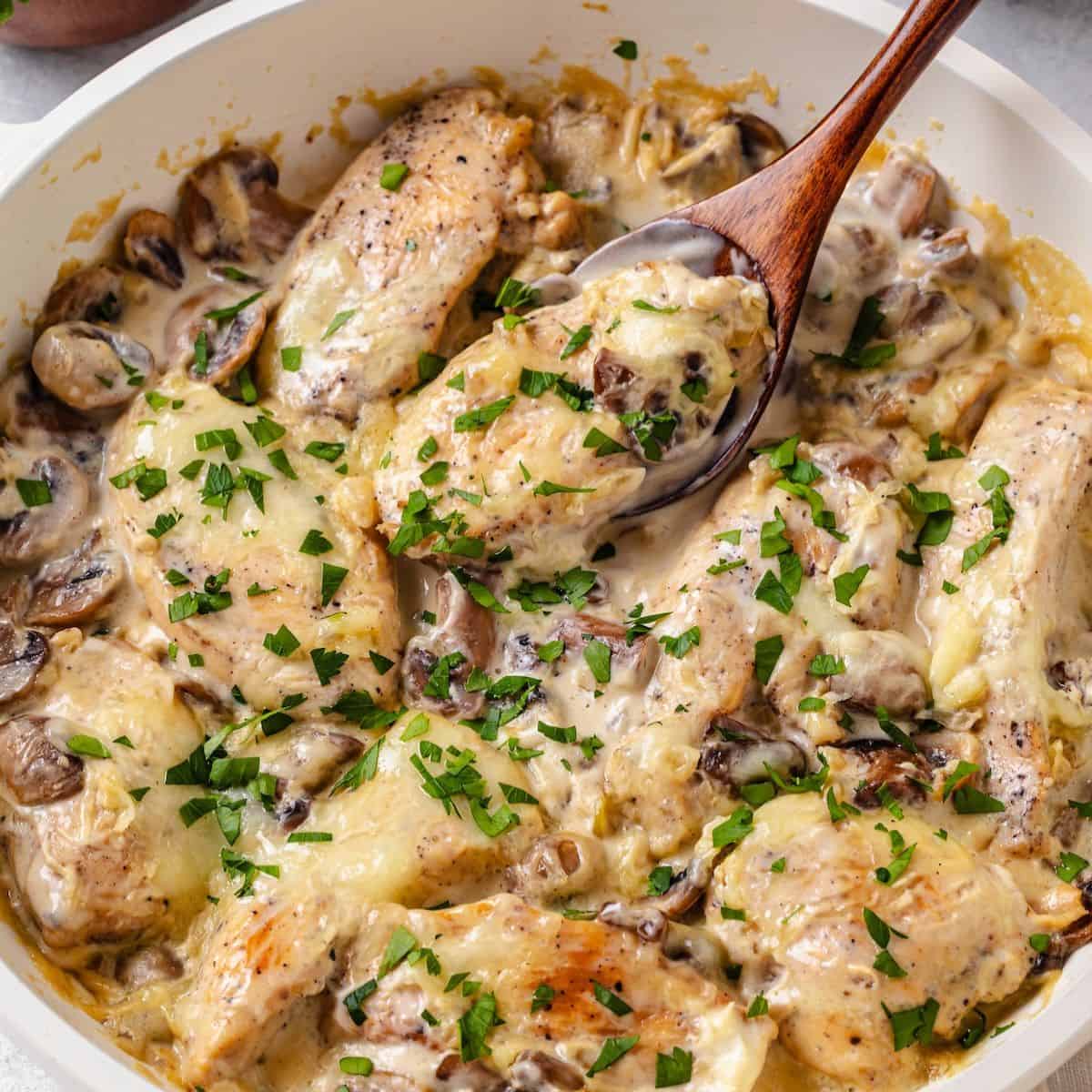 Close up of a spoon scooping up a piece of chicken from a skillet full of chicken in a cream cheese sauce with parsley sprinkled on top.