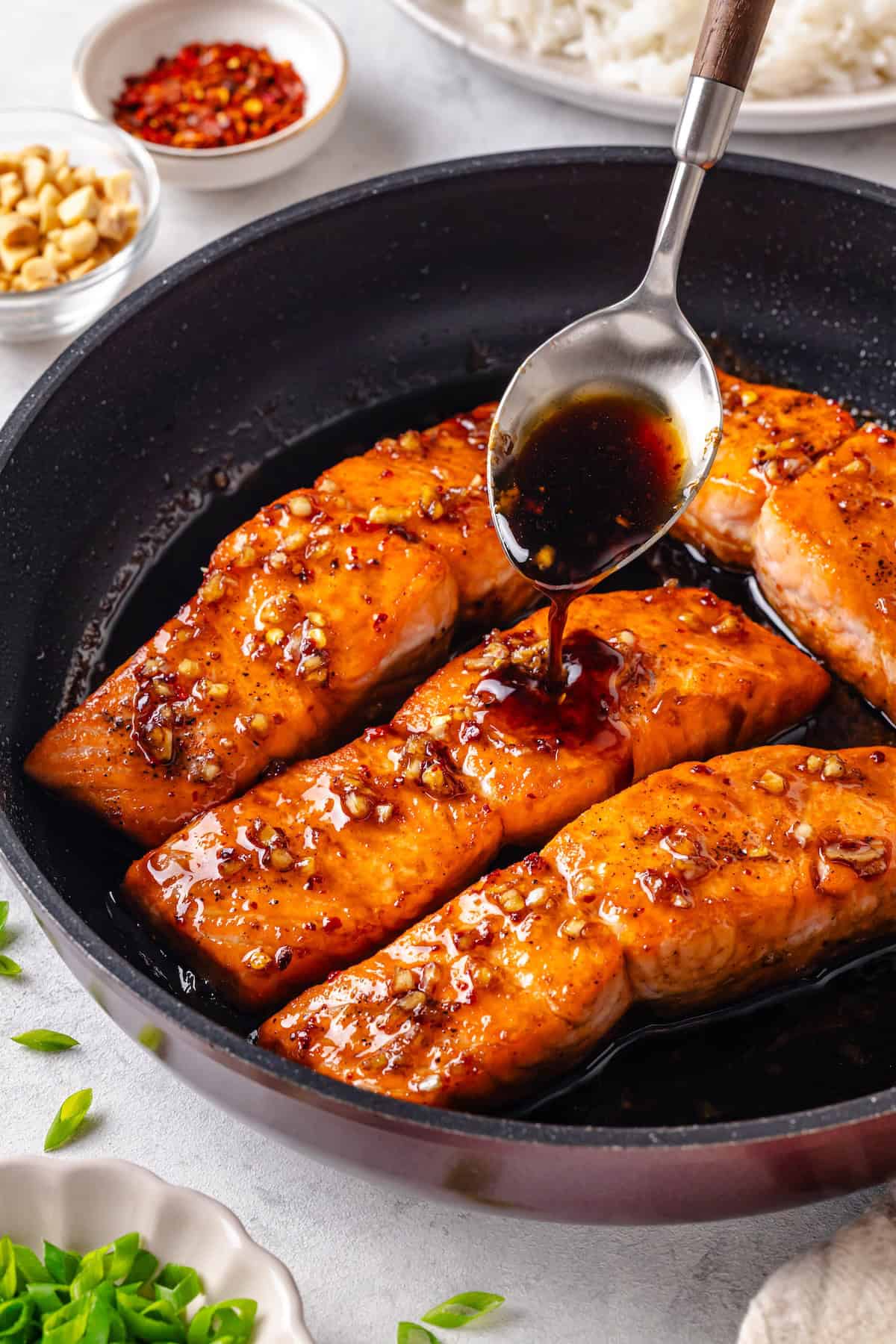 A spoon drizzling a honey garlic sauce over salmon filets in a dark skillet.