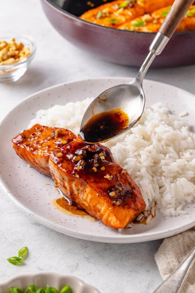 A spoon drizzling honey garlic sauce over a piece of salmon on a plate with some steamed white rice.