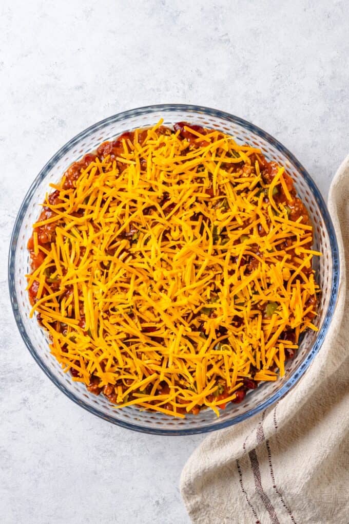 Shredded cheddar cheese spread on top of a layered Mexican dip in a glass pie plate before being baked.