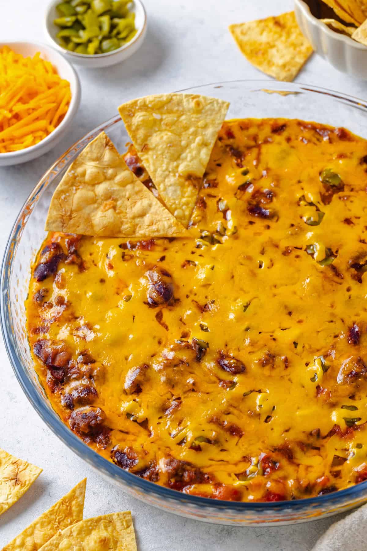 A baked chili dip appetizer with melted cheese on top in a glass pie plate with chips resting on the side.