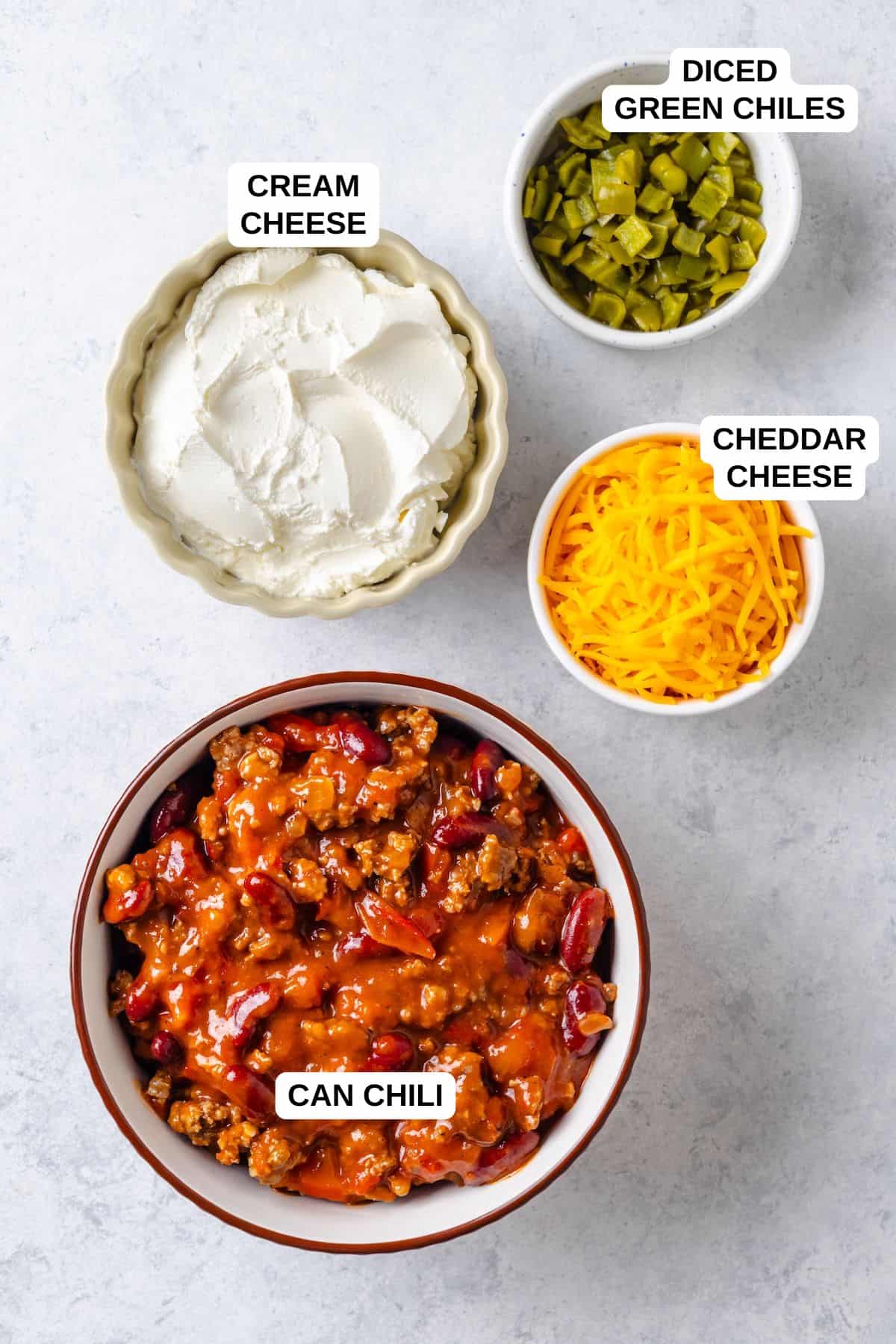 Four ingredients laid out in separate bowls for making a Mexican dip, including canned chili, cream cheese, green chiles and cheddar cheese.