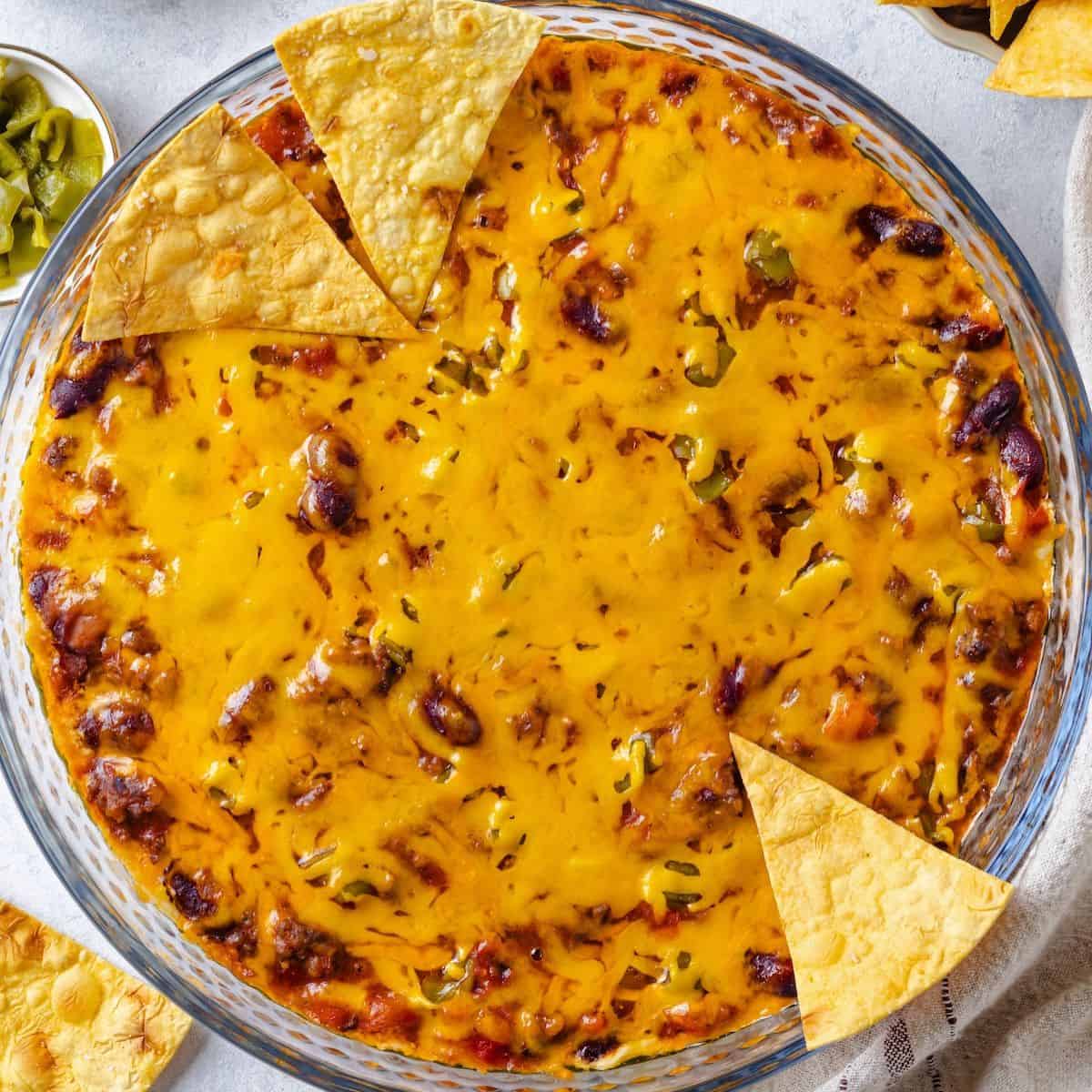 Close up of chips resting on the sides of a glass pie plate with a cheesy baked Mexican dip with canned chili.