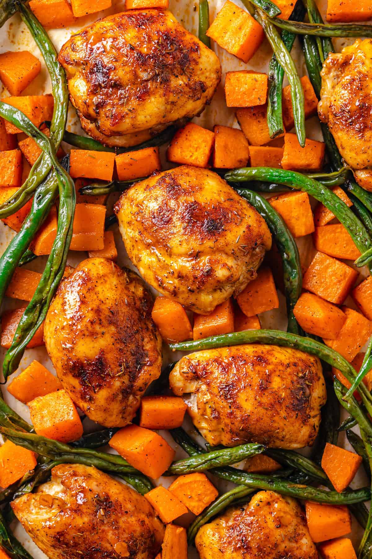 Close up of chicken thighs with sweet potatoes and green beans on a sheet pan after being cooked.