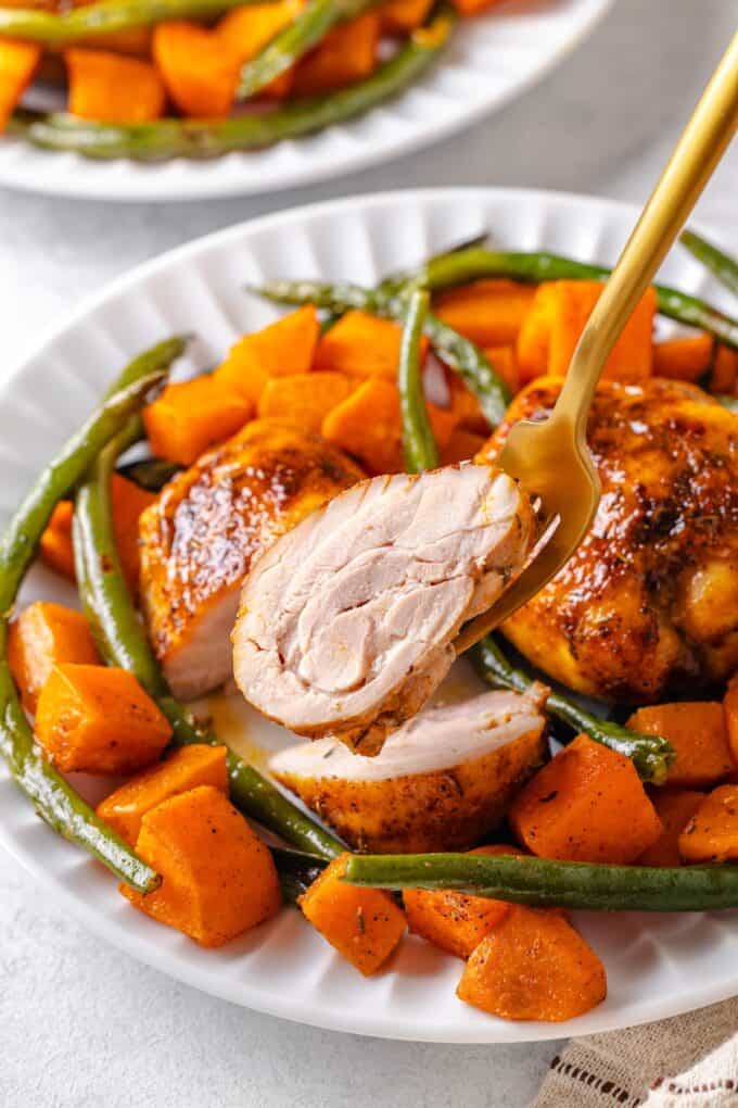 A fork lifting up a slice of a baked chicken thigh from a white dinner plate.
