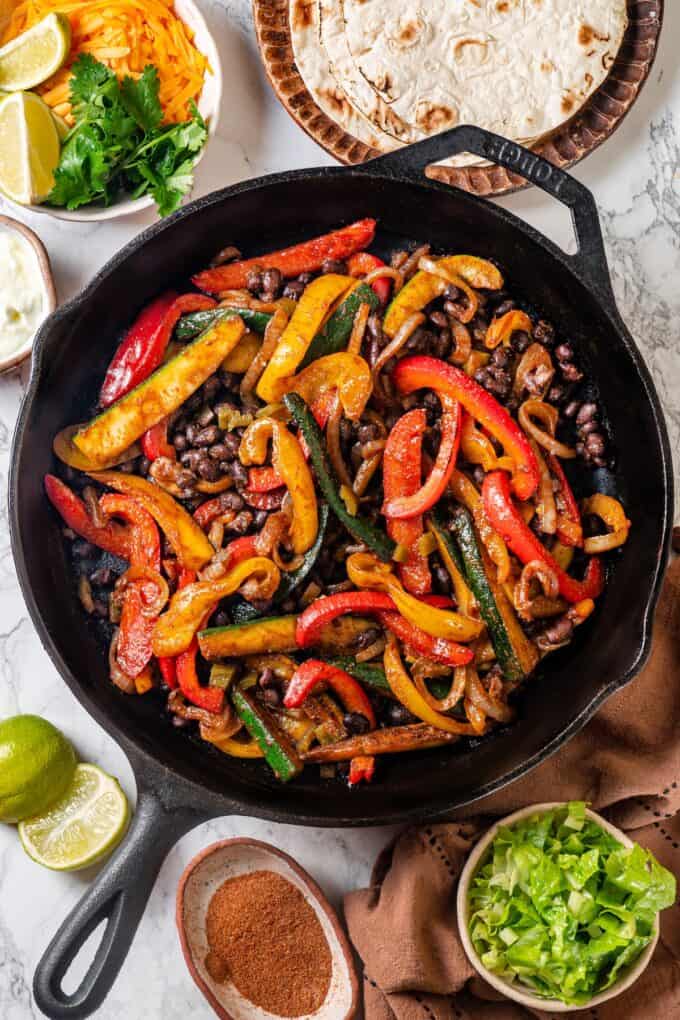Black bean and veggie fajita skillet in a cast iron skillet with tortillas and toppings to the side.