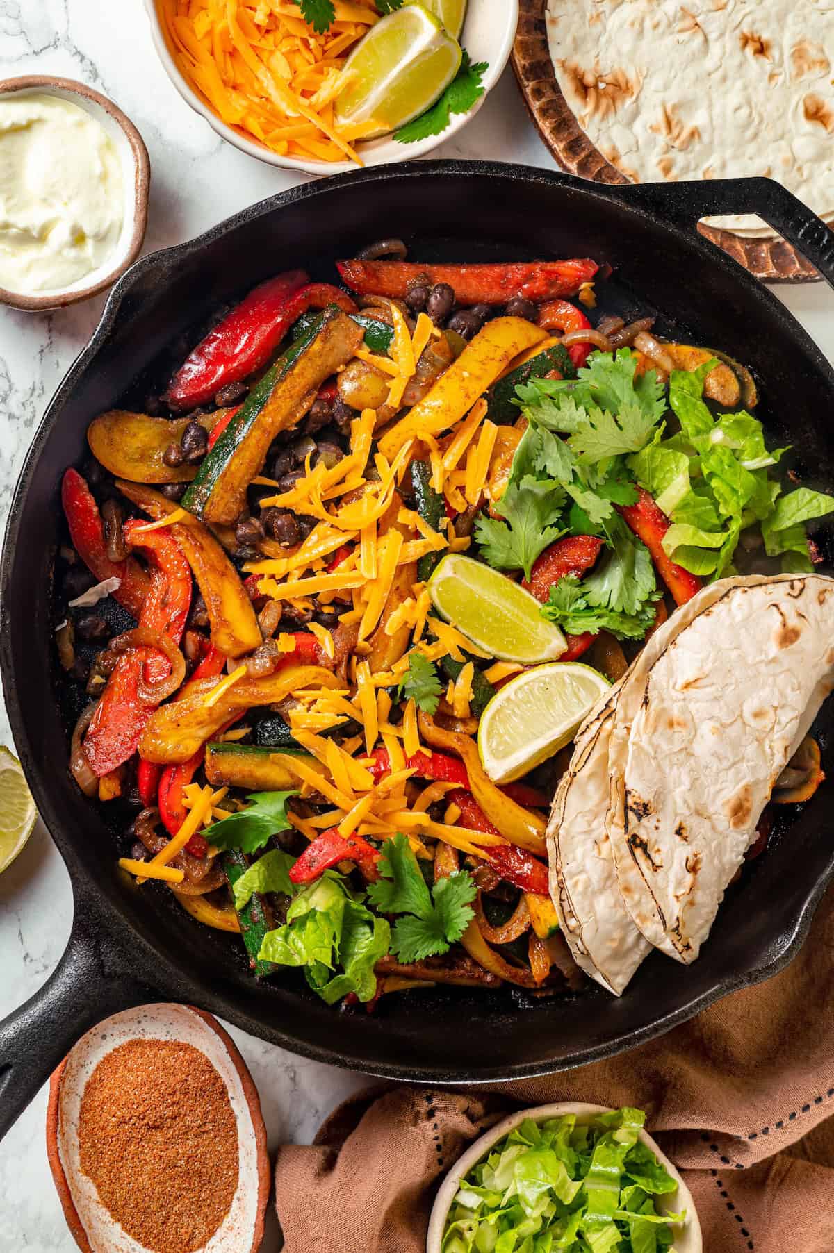 A vegetarian fajitas skillet with peppers, zucchini and black beans in a cast iron pan with charred tortillas to the side.
