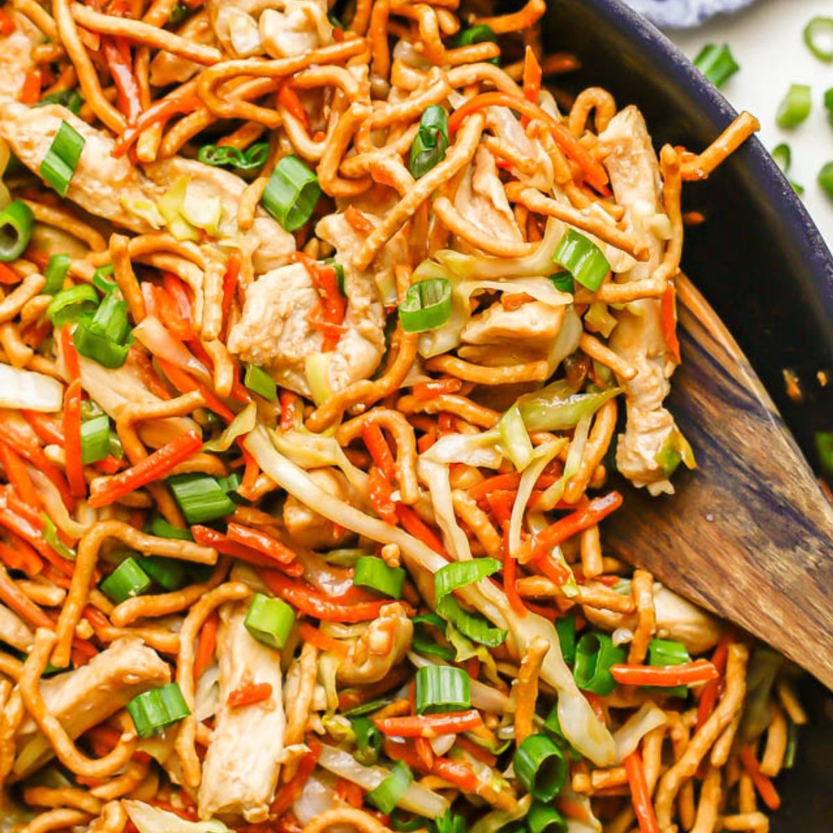 Close up of a wooden spatula in a skillet with a chicken chow mein mixture with green onions sprinkled on top and to the side.
