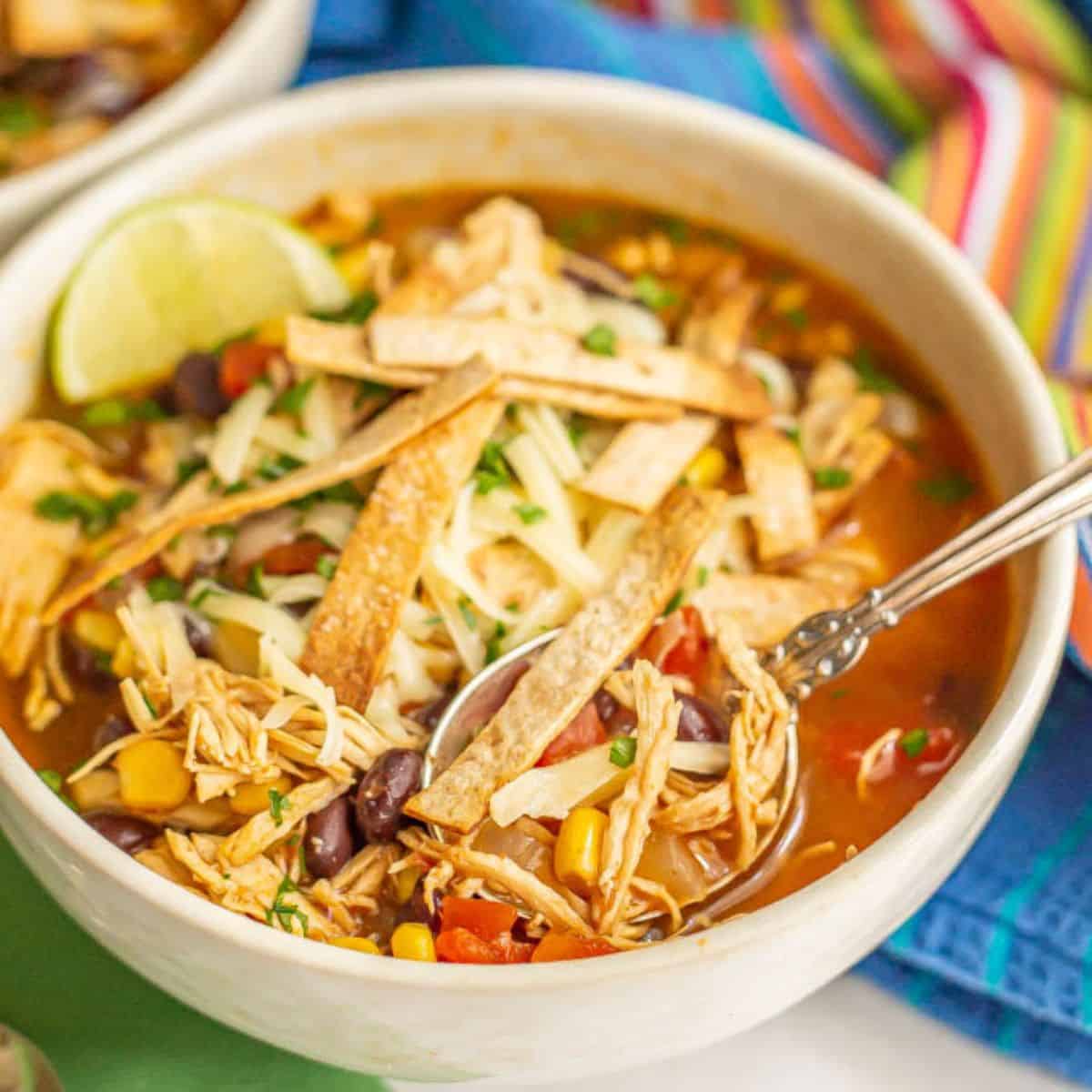 Close up of a bowl of chicken tortilla soup with black beans and corn and crisp homemade tortilla strips on top and a spoon resting in the bowl.