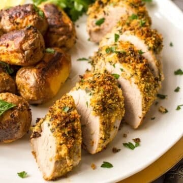 Close up of an herb crusted pork tenderloin sliced and served on a white platter with roasted potatoes to the side.