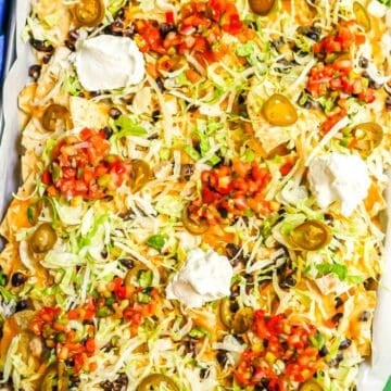 Close up of loaded chicken and black bean nachos on a parchment paper lined sheet pan with pico de gallo, sour cream, pickled jalapeños and shredded lettuce on top.