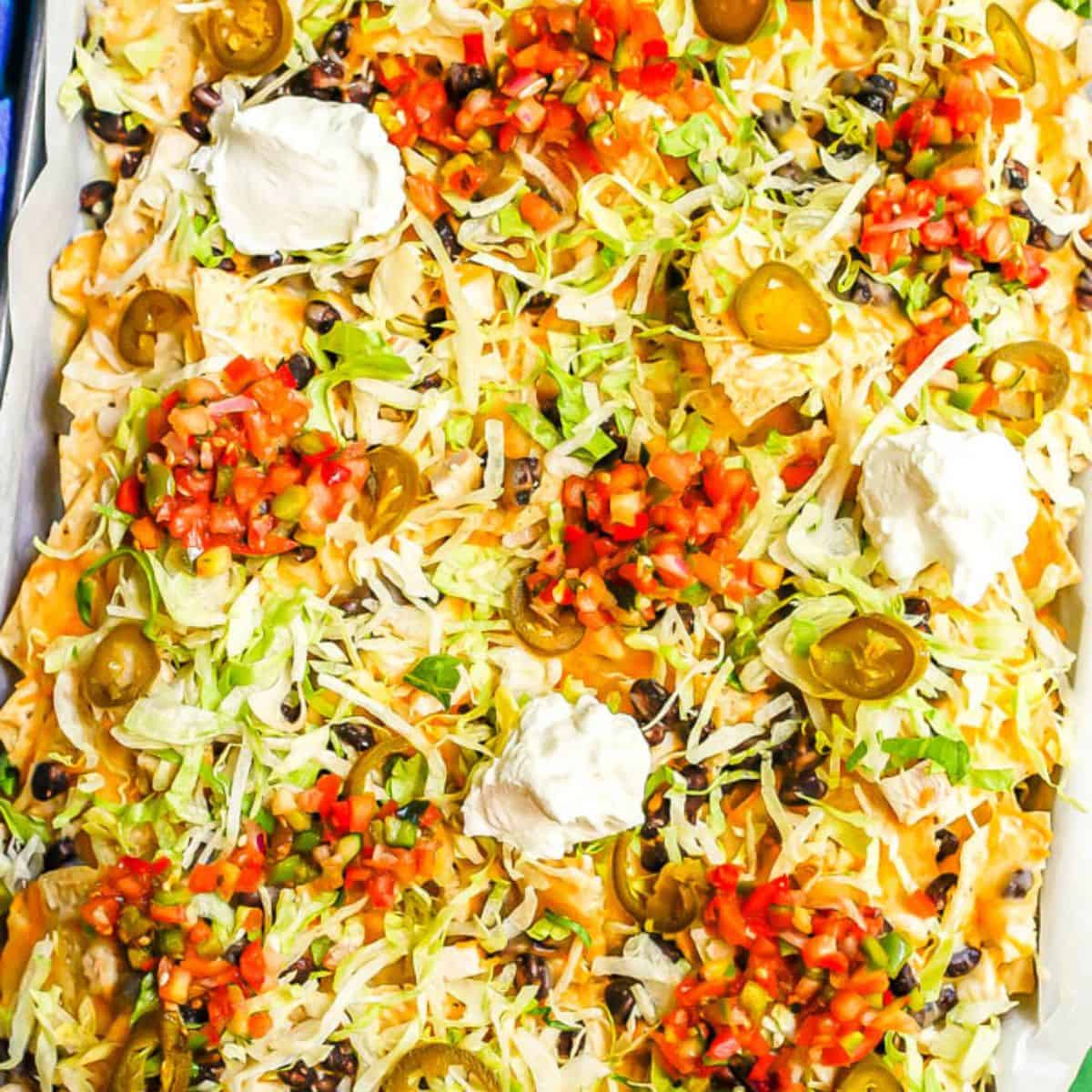 Close up of loaded chicken and black bean nachos on a parchment paper lined sheet pan with pico de gallo, sour cream, pickled jalapeños and shredded lettuce on top.