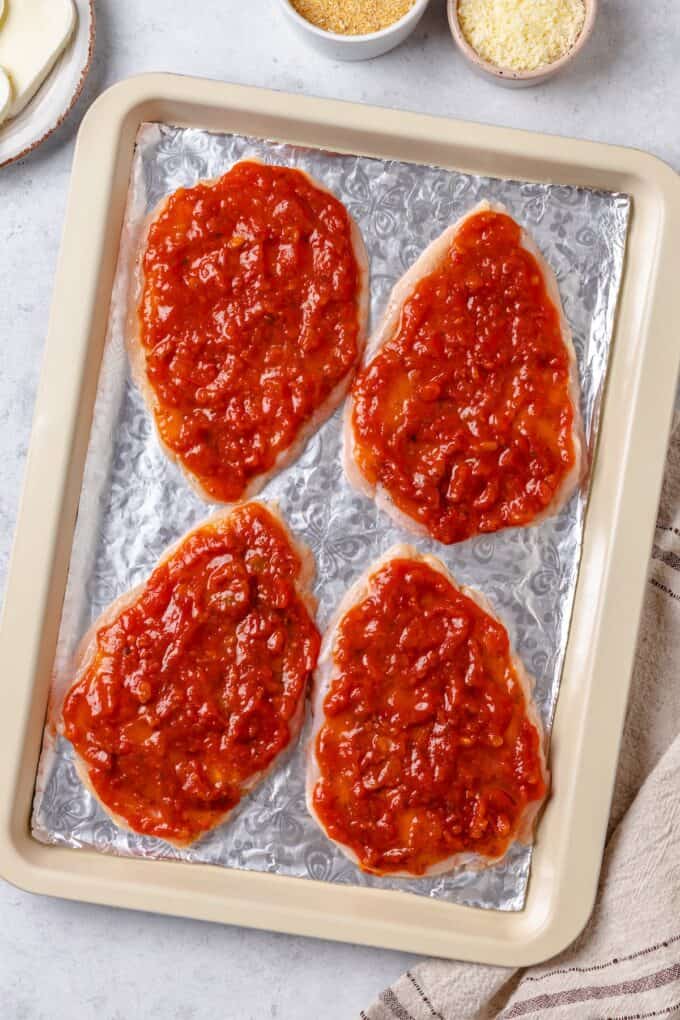 Chicken breasts topped with marinara sauce on a foil lined baking sheet.