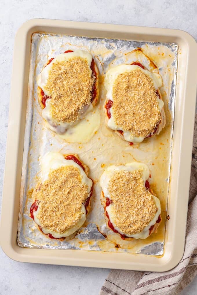 Breadcrumbs on top of melted mozzarella cheese and marinara on top of chicken breasts on a foil lined sheet pan.