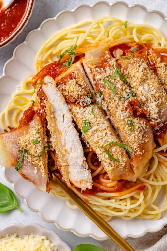 Sliced chicken Parm served over spaghetti noodles and marinara sauce on a scalloped dinner plate.