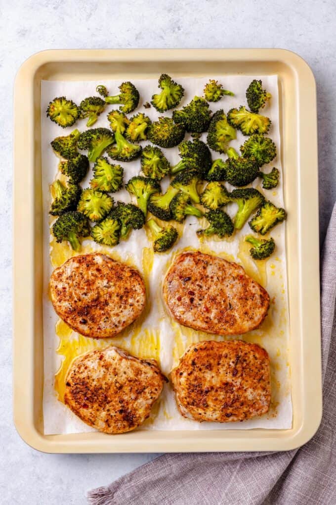 Cooked pork chops on a parchment paper lined baking sheet with roasted broccoli on the other side.