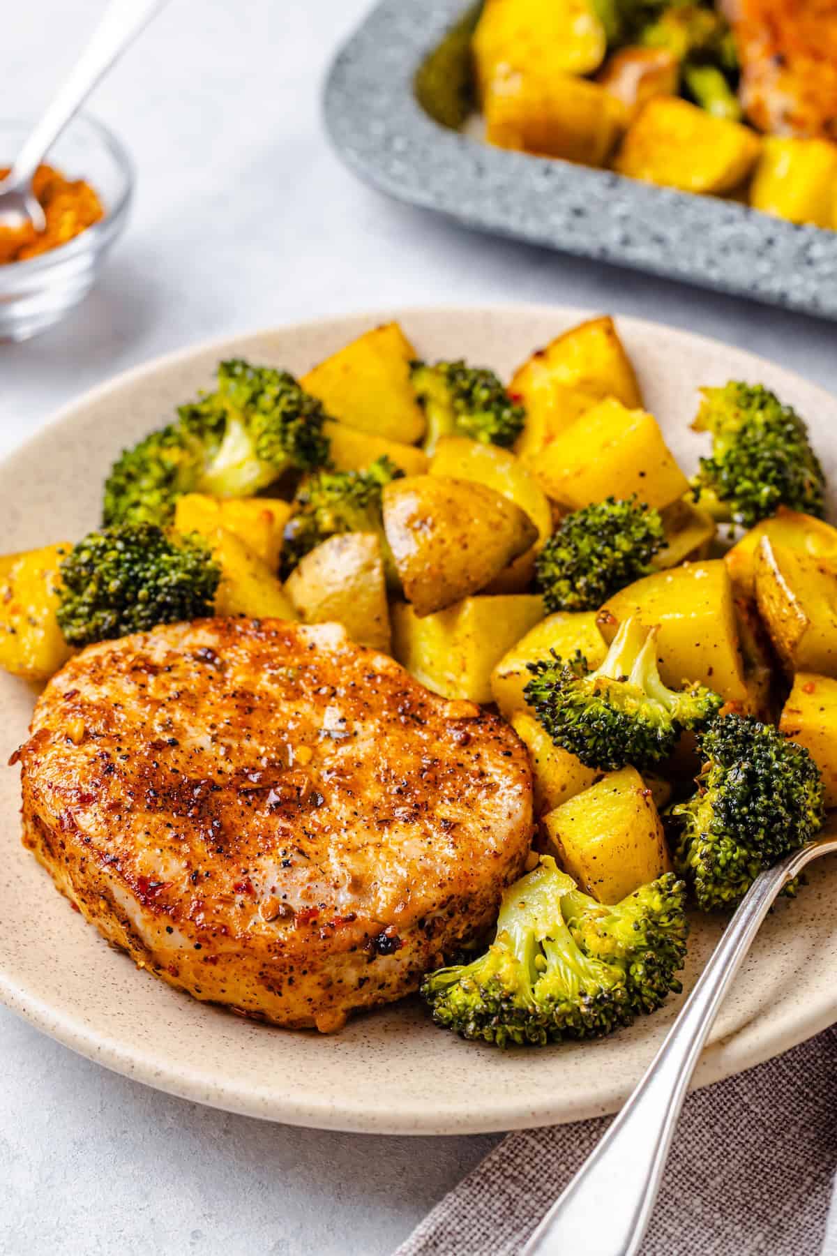 A dinner plate with a Cajun seasoned roasted pork chop alongside a mixture of roasted potatoes and broccoli with a fork to the side.