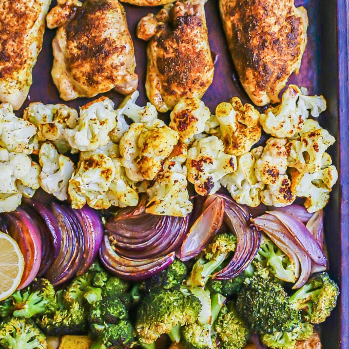 Close up of a sheet pan dinner with chicken thighs, roasted cauliflower, red onion slices and broccoli arranged in rows.