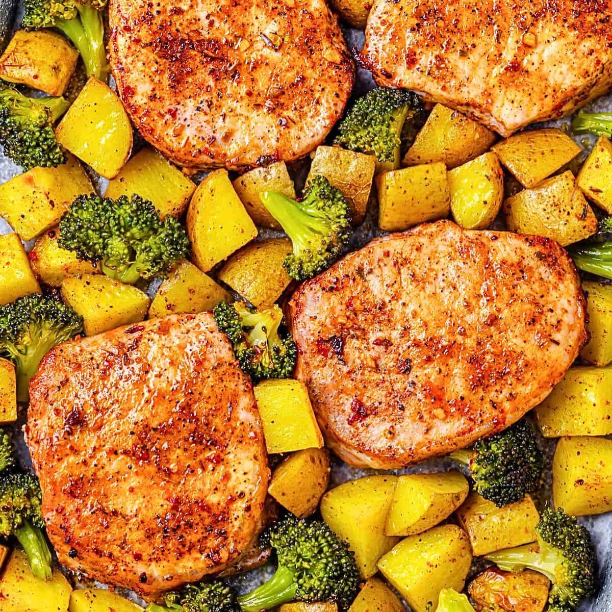 Close up of pork chops nestled in among roasted potatoes and broccoli on a sheet pan.