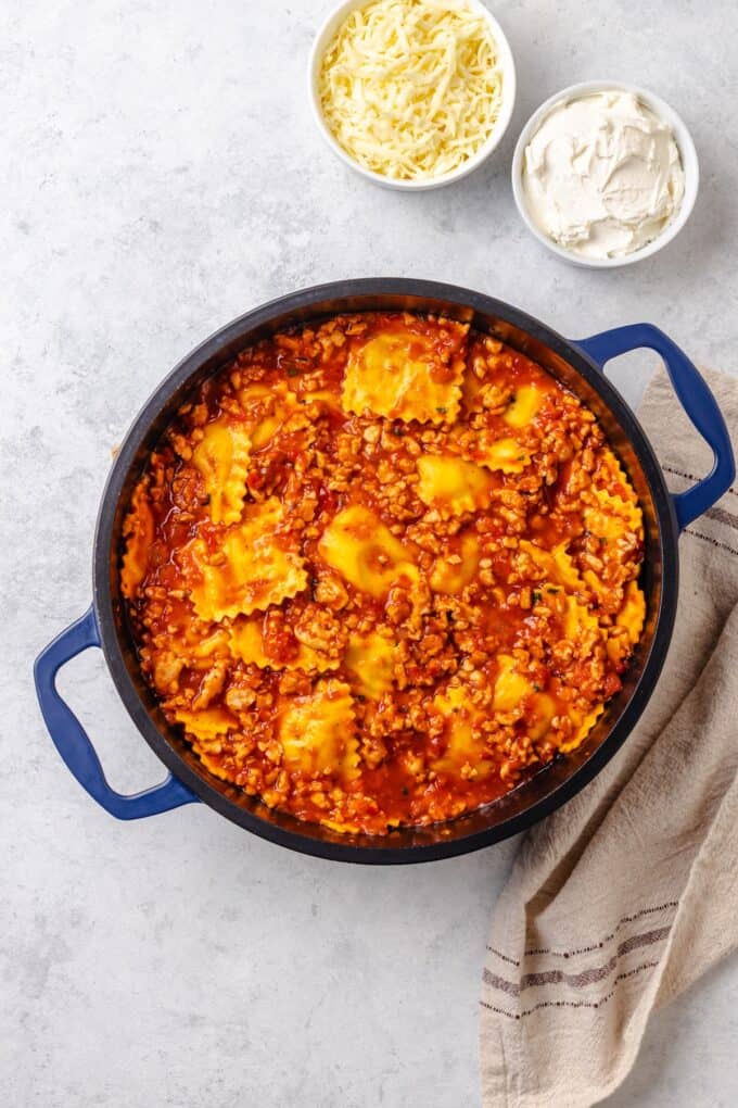 A large blue pot with a saucy ground turkey and ravioli mixture with two bowls of cheese to the side before being added in.
