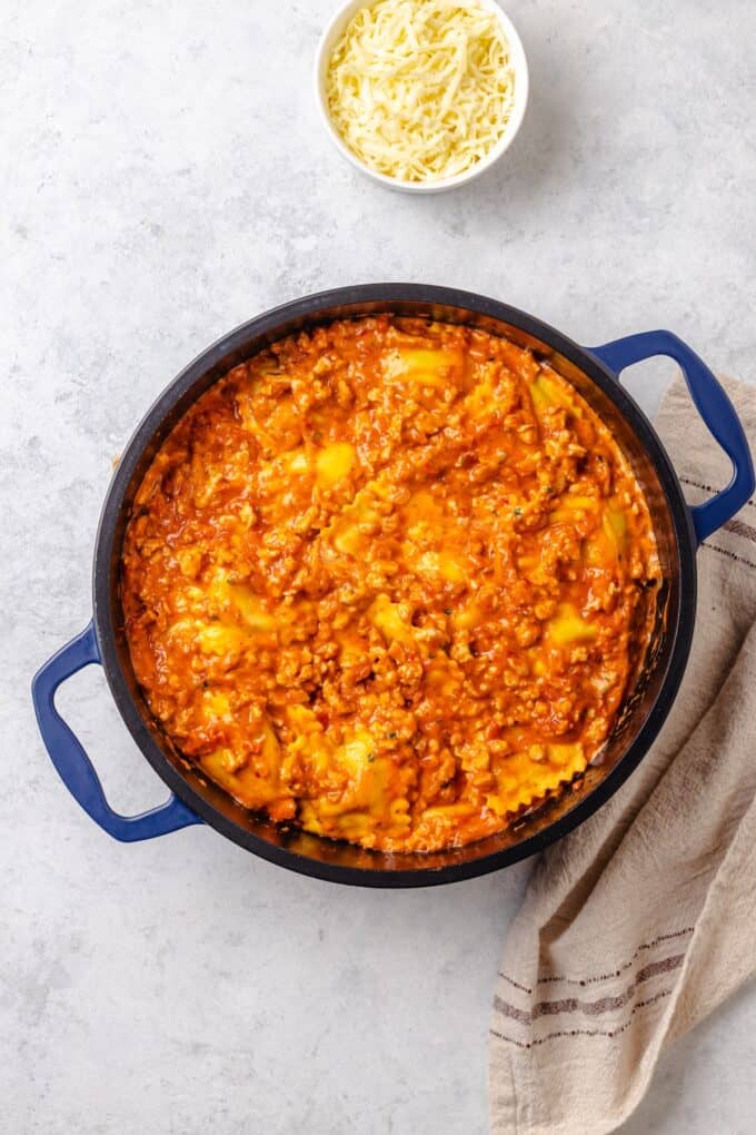A creamy tomato and ground turkey mixture with ravioli in a large blue pot with a bowl of mozzarella cheese to the side.