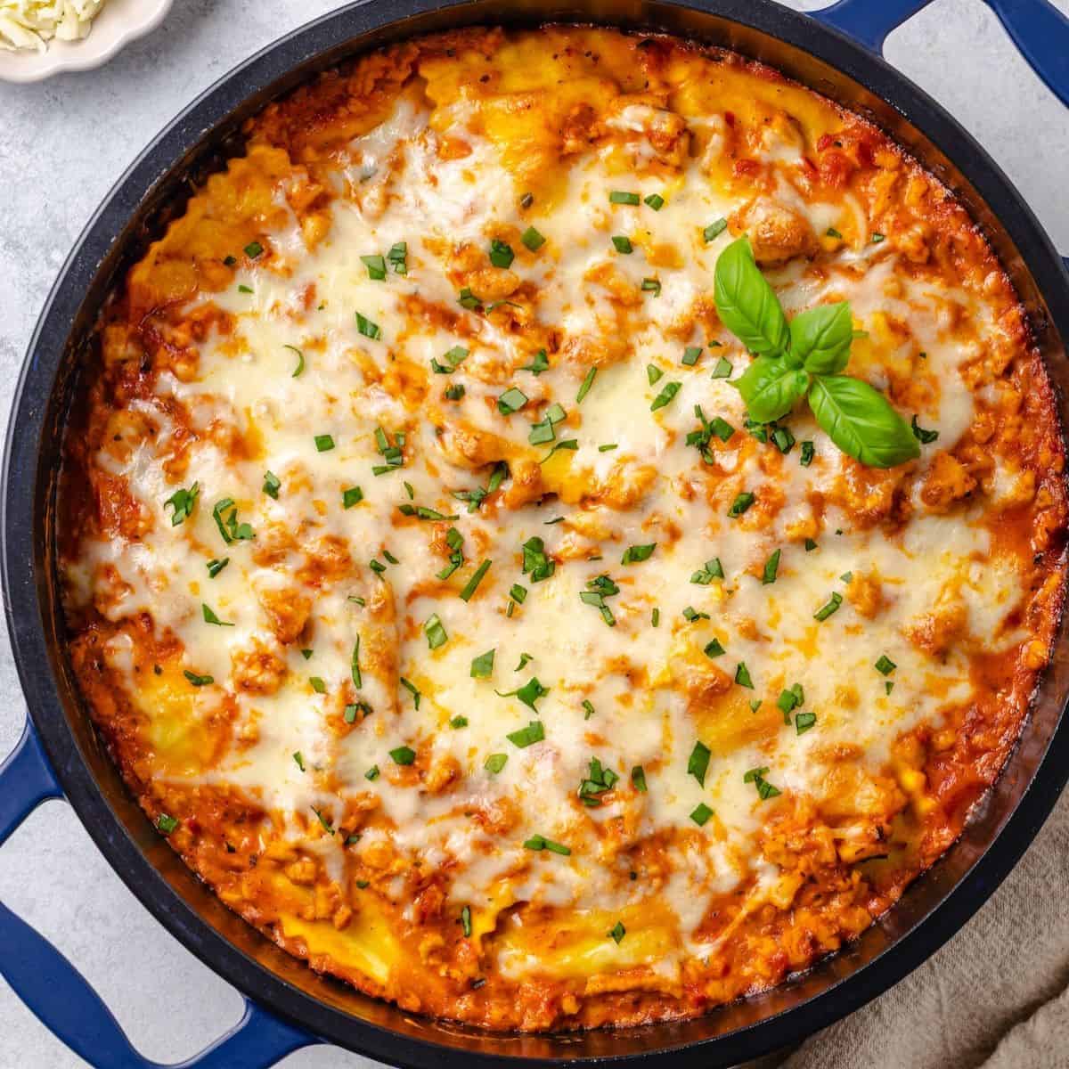 Close up of a ravioli lasagna in a deep blue skillet with melted mozzarella and fresh basil on top.