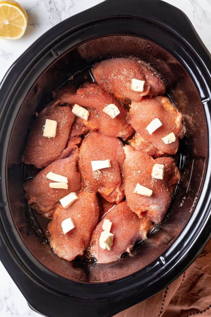 Seasoned chicken thighs topped with small pats of butter in a slow cooker before being cooked.