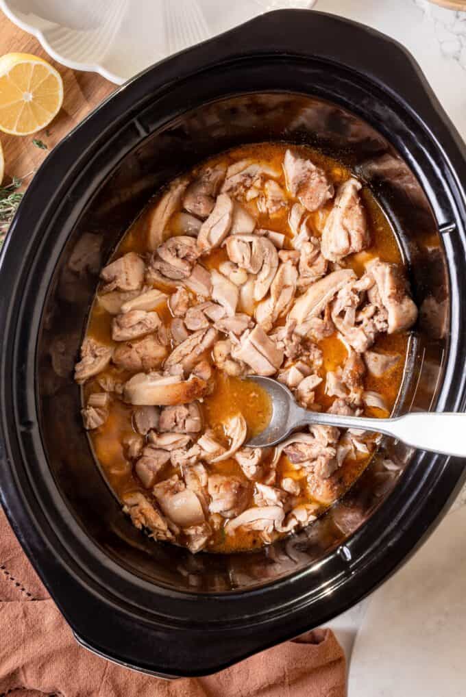 A slow cooker with cooked, chopped chicken thighs being stirred into the cooking juices and sauce.