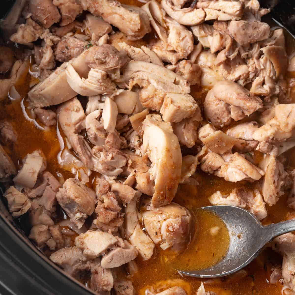 Close up of a spoon resting in a slow cooker with cooked, chopped chicken thighs in a brothy sauce.