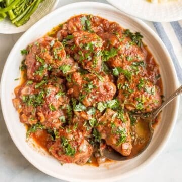 Close up of a round white serving dish filled with saucy French style slow cooker chicken thighs with fresh parsley on top and a serving spoon resting in it.