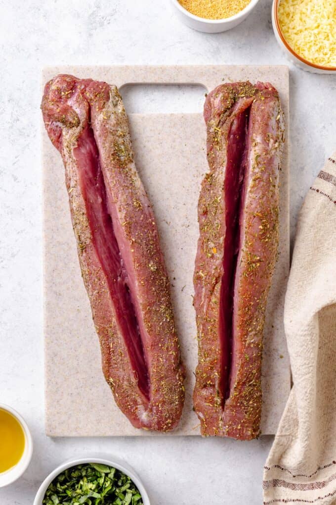 Two pork tenderloins on a cutting board with a large slit cut into the tops of each one for stuffing.
