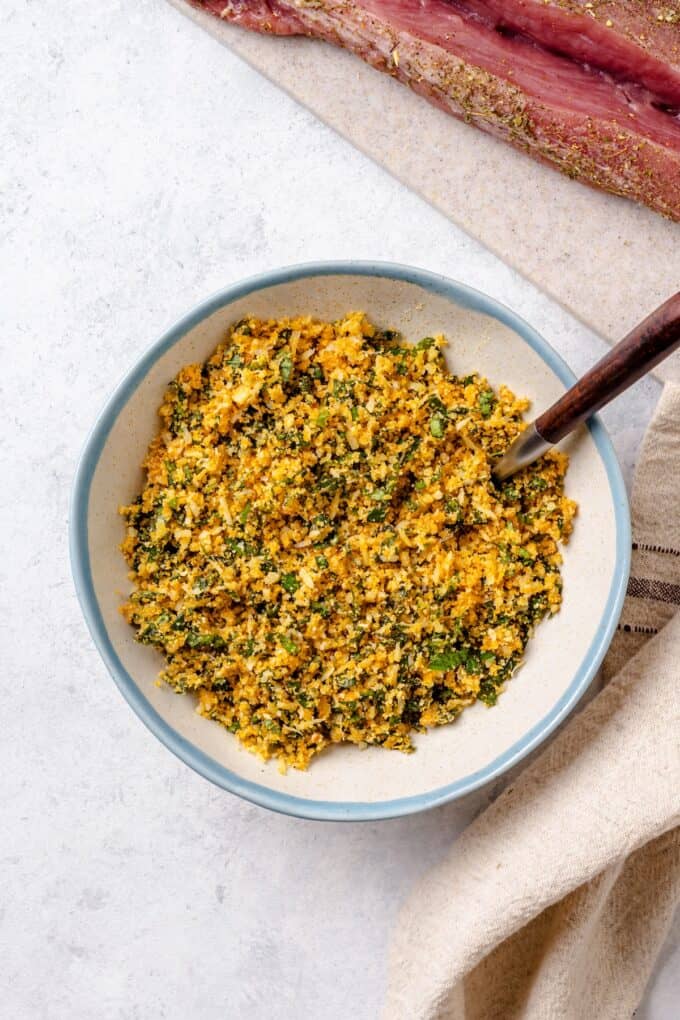 A small bowl with a breadcrumb, Parmesan cheese and basil mixture and a spoon resting in it.