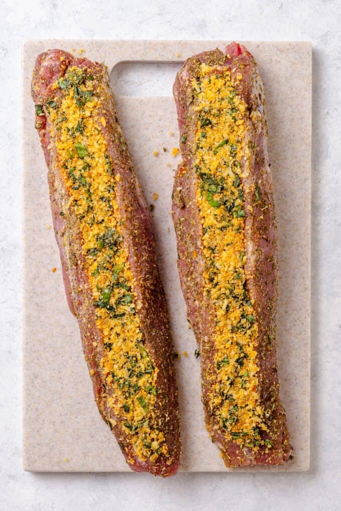 Two pork tenderloins on a cutting board with a breadcrumb and basil stuffing mixture added to each one.