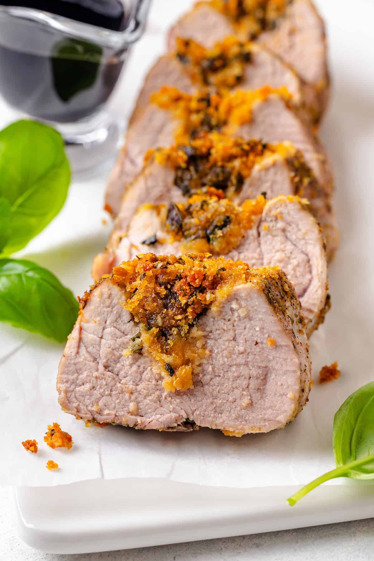 Sliced stuffed pork tenderloin on a serving tray with fresh basil to the side and a container of balsamic glaze in the background.