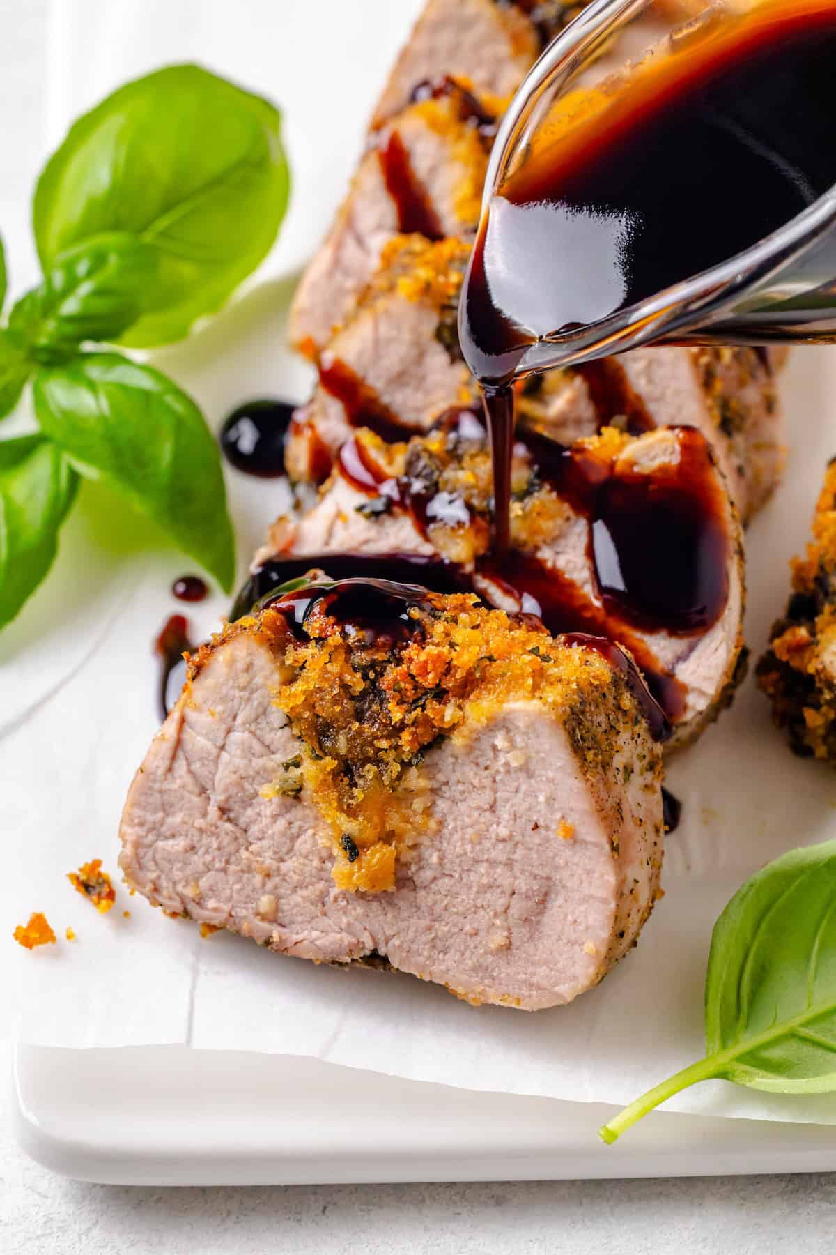 Balsamic glaze being poured over sliced stuffed pork tenderloin on a serving tray with fresh basil to the side.