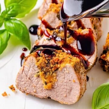 Close up of balsamic glaze being poured over sliced stuffed pork tenderloin on a serving tray with fresh basil to the side.