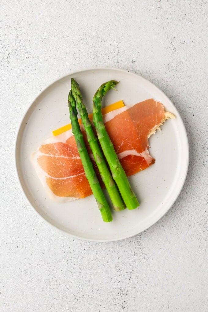 A plate with a crescent roll dough triangle topped with a thin slice of cheddar cheese, prosciutto and three asparagus spears.